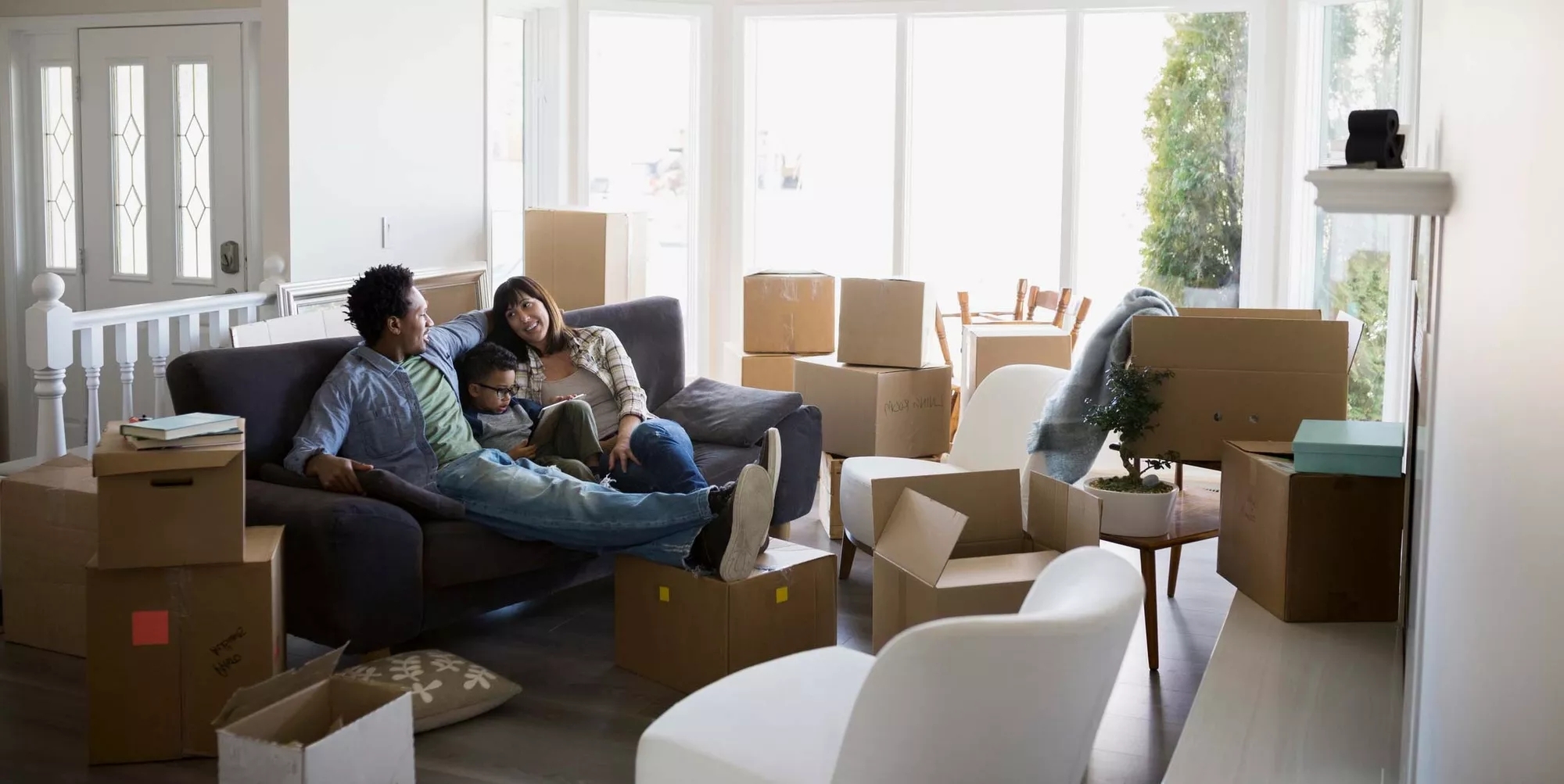A smiling family resting on a couch, in a living room crowded with moving boxes. They appear to be moving in.